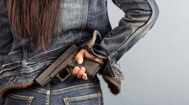 Woman wearing blue denim jacket and pants, viewed from the rear, where we can see she's concealing a semi-automatic handgun.